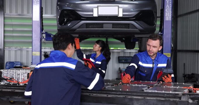 Lead mechanic and technician discuss electric vehicle battery testing using a handheld diagnostic meter while another mechanic inspects a raised car. 