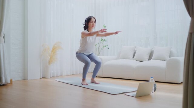 Young Asian beautiful woman doing aerobic exercise at home. Attractive girl doing  workout by squatting, follow instructions video from online trainer for health in house