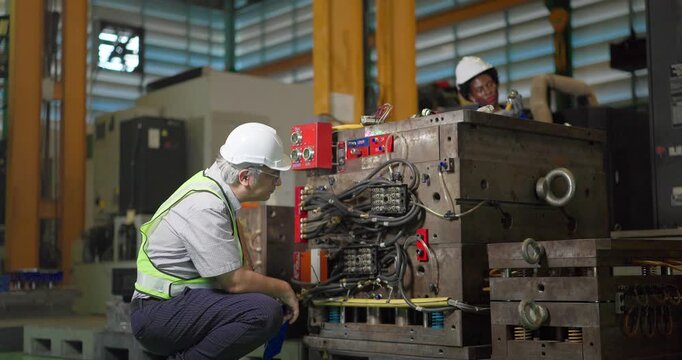 A senior male engineer in a safety vest and hard hat crouches beside a large steel injection mould, checking its hydraulic hoses, manifold blocks and wiring and writing notes on a clipboard.
