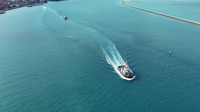 tugboats sailing through clear blue sea water near a concrete breakwater. Maritime logistics support, harbor navigation, and commercial port operations concept.