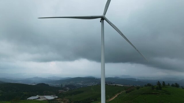 Panning shot of wind turbine rotating over green Cau Dat Tea Hill under cloudy sky Da Lat, Vietnam 