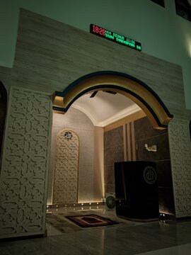 Interior view of a mosque featuring a beautifully decorated mihrab with Islamic geometric patterns, an archway, a prayer rug, a wooden minbar, and a digital prayer time clock on the wall.