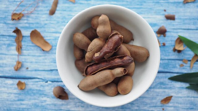 Fresh tamarind fruits in white bowl on blue wooden background with dry leaves motion.