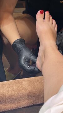 A close-up of a woman's foot with red nail polish being pampered in a spa. A gloved hand is applying treatment to the ankle area.