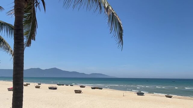 A serene beach scene with golden sand, clear blue water, and distant mountains. Palm trees frame the view, creating a tropical atmosphere.