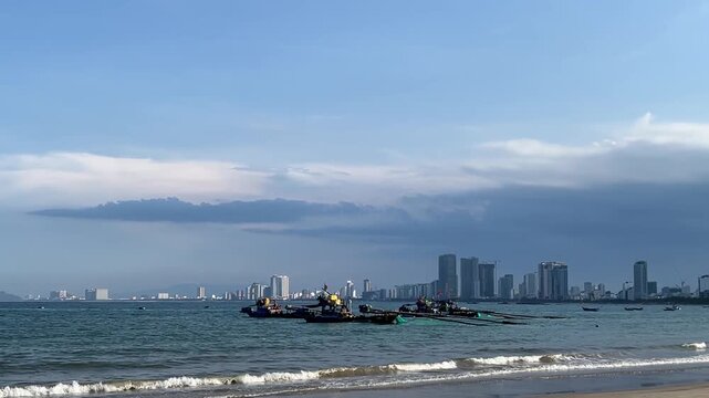 Fishing boats on a calm sea near a city skyline. The sky is partly cloudy with a blue hue. Waves gently lap at the shore.