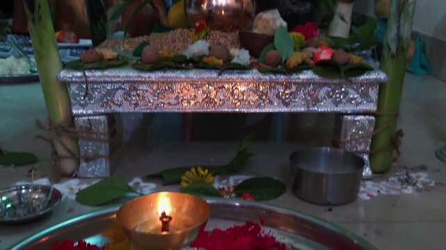 Silver altar table holding a copper kalash with coconut over wheat grains, betel nuts, and flowers next to a burning camphor flame on a steel pooja thali during a traditional house ceremony.