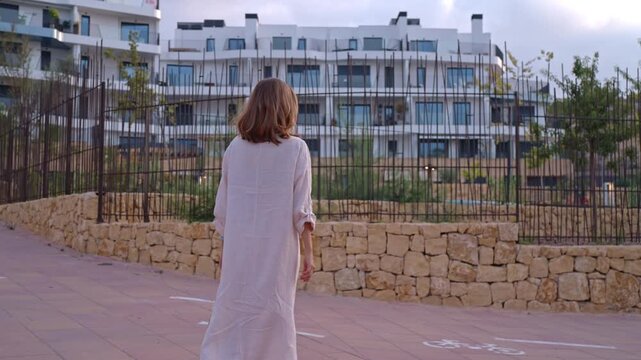 A person walks along a path in a residential area during the late afternoon. The buildings have many windows, and the sky has some clouds.