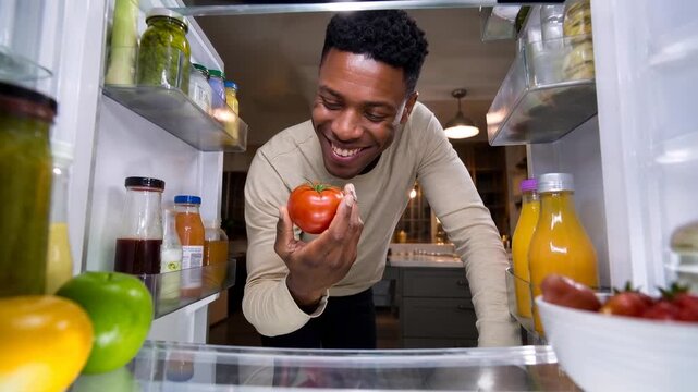 Man happily selecting fresh tomato from a well-stocked refrigerator. A smiling man reaches into a brightly lit refrigerator filled with various food items and drinks to pick out a