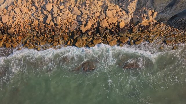 This shows waves hitting rocks along a beach during sunset. The sun casts shadows over the water and the rocky shore. The scene captures nature's beauty at dusk.