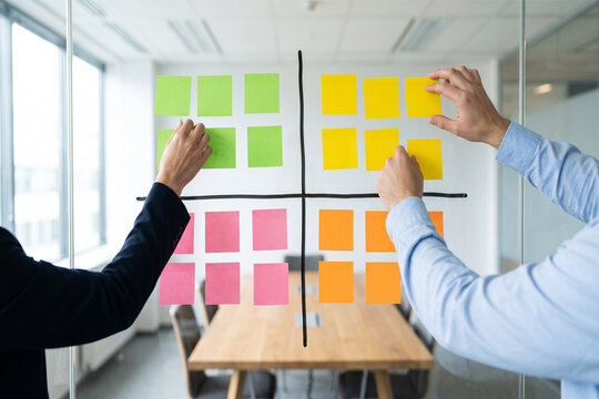 Colleagues organizing tasks on a quadrant priority matrix with sticky notes on a glass wall to prioritize projects, improve workflow and support agile team planning in a modern office