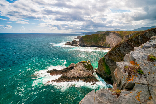 Breathtaking rugged coastline with turquoise ocean waves crashing against limestone cliffs and a rocky sea stack beneath a dramatic cloudy sky, evoking wild, untamed coastal beauty and adventure.