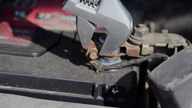 a mechanic using an adjustable wrench to loosen a rusty bolt on a car battery terminal under the hood.