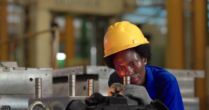 A Black female technician in a hard hat leans over a workbench and holds a small metal component close to her eye to inspect it beside a large aluminium injection mould in a toolroom.
