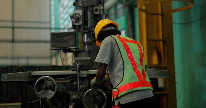 A Black female machinist in a hi vis vest and hard hat turns the handwheels of a knee type milling machine to feed the table while cutting a metal part in a factory workshop.
