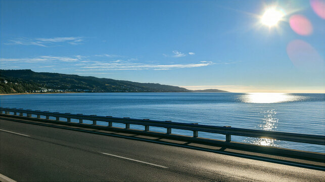 Eye level side view of empty asphalt road beside coastal water, with sunlight, mountain shoreline, guardrail, and clear blue sky creating peaceful scenic landscape