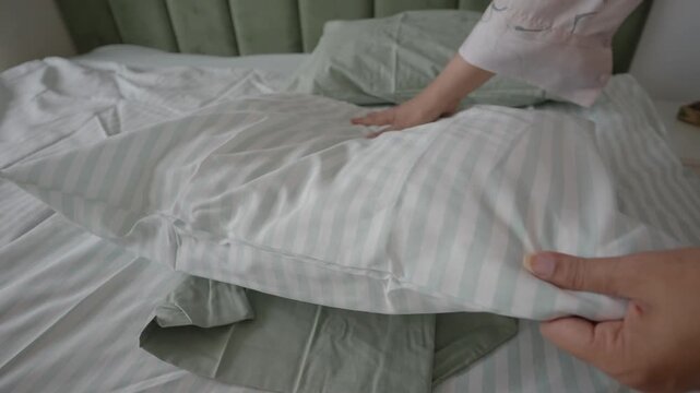 Woman hands smoothing striped pillow on unmade bed while tidying up bedroom in morning sun.