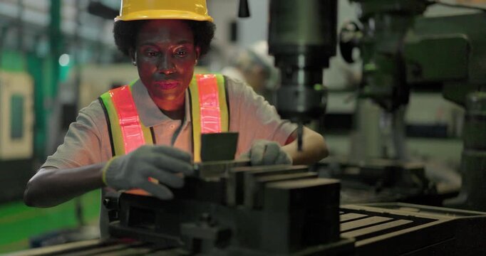 A Black female machinist in a hi vis vest, hard hat and safety glasses sets a metal workpiece in the machine vice of an industrial drilling machine and operates the spindle in a factory.
