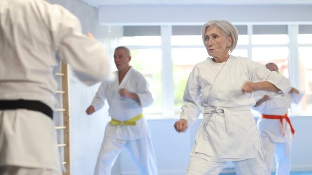Diligent old man attendee of karate classes practicing kata standing in row with others in sports hall 