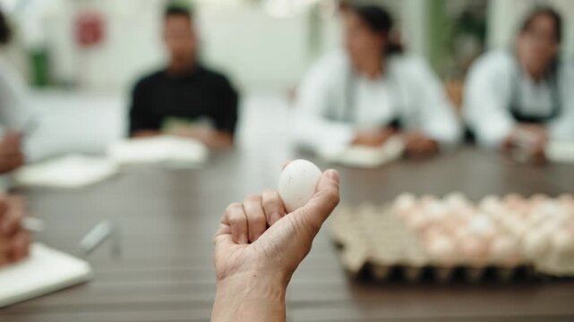 Hand holds a single white egg above a tray of eggs while people listen in the blurred background of a food workshop.