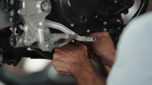 Hands of a mechanic reach under the crankcase of a motorcycle to work on the footpeg and engine casing in a repair shop.