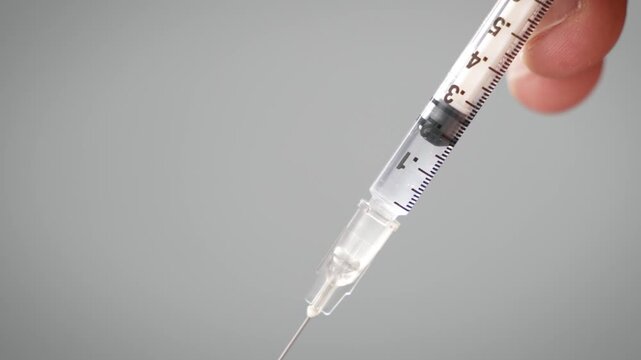 Locked off close up of liquid being pushed out of a syringe held in front of a plain grey background [TRT Concept]