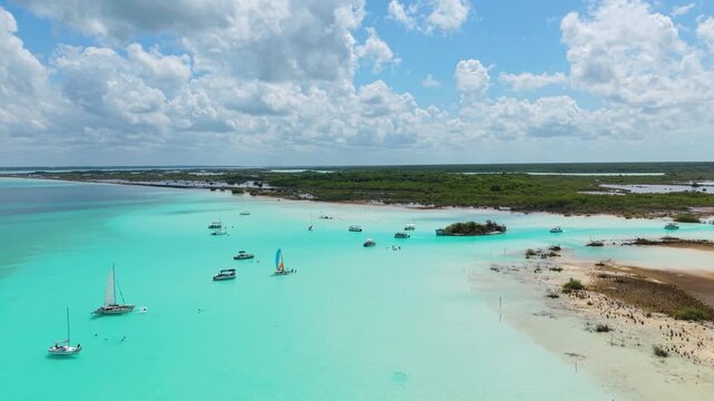 Sailboats and motorboats float across crystal-clear aquamarine shallows near a forested shore.