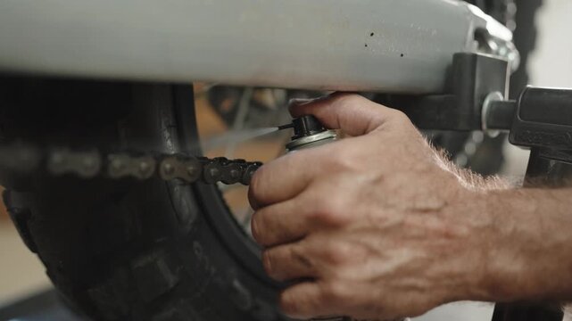 Hand of a mechanic sprays lubricant along the drive chain of a motorcycle raised on a stand in a repair shop.