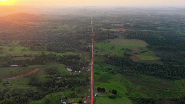 Red-soil agricultural fields, pasture and scattered woodland along a long rural road near Nueva Alborada, Itap&uacute;a, Paraguay, with Paran&aacute; basin countryside under humid skies, drone forward flyover