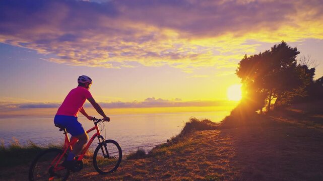 A senior adult, dressed in vibrant cycling gear, stands beside his red bike as he gazes at the breathtaking sunset over a serene lake.