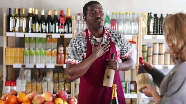 In store, African male seller communicates with woman buyer and offers to buy wine, shows assortment, gives bottle of red wine to choose. Store employee serves customer. 