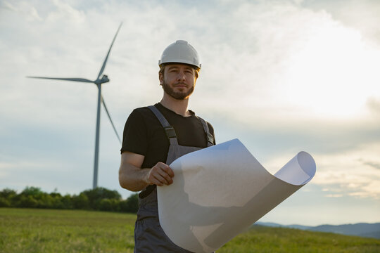 A Caucasian man in work clothes and hard hat stands in a grassy field with a wind turbine behind him, holding rolled-up plans