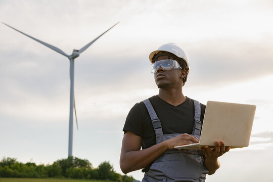 A man in a hard hat and safety glasses holds a laptop while standing in a field with a wind turbine in the background