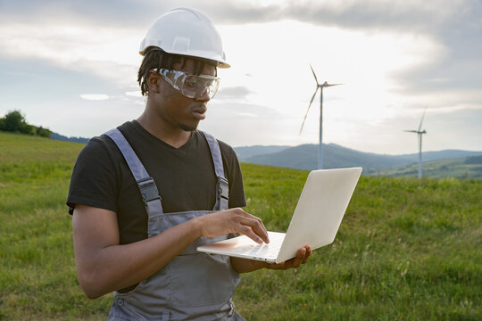 A focused African American man wearing protective gear works on a laptop amidst a grassy field with wind turbines in the background
