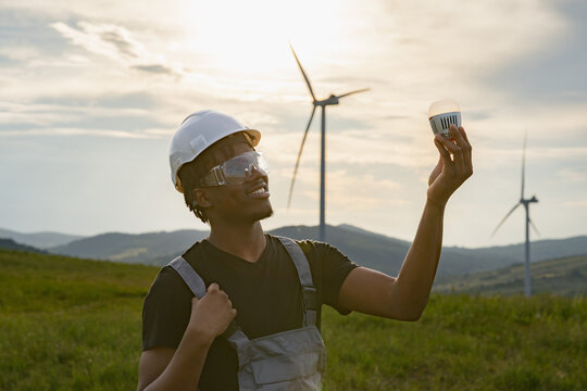 A smiling African American man in a hard hat and safety glasses holds an LED light bulb while standing in a field with wind turbines