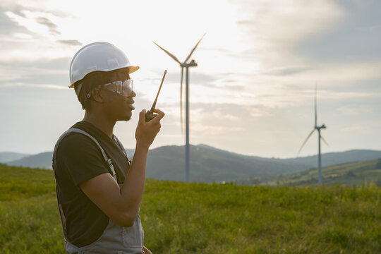 A man wearing protective gear communicates on a radio while standing in a grassy field with wind turbines in the background