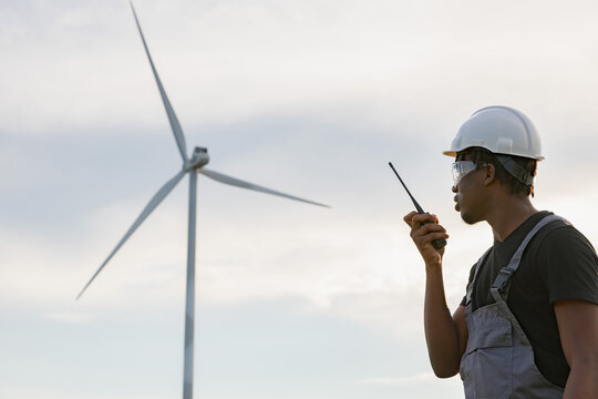 A man in a hard hat and overalls speaks into a walkie-talkie with a wind turbine in the background