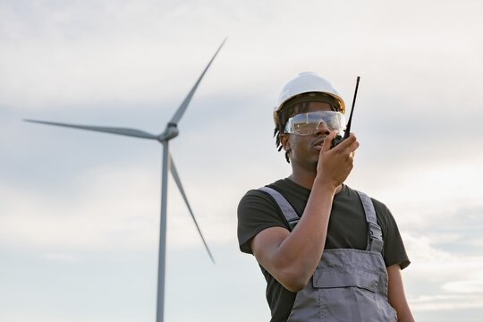 A focused African American man in safety gear speaks into a walkie-talkie with a wind turbine in the background