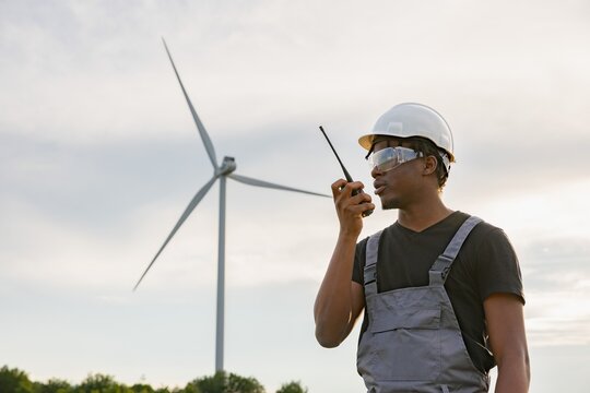 A man in work clothes and safety gear speaks into a walkie-talkie with a wind turbine in the background, suggesting renewable energy operations