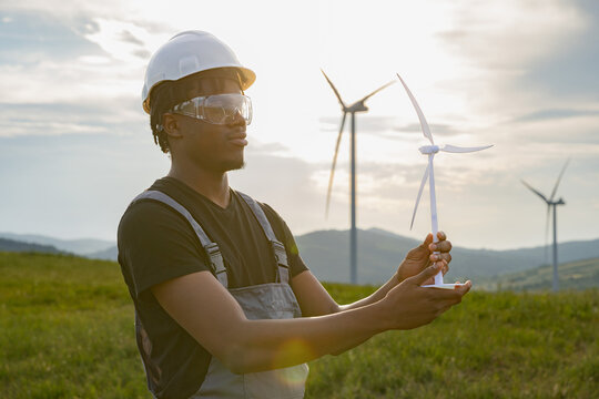 A young African American man in safety gear holds a miniature wind turbine model in a field with large wind turbines behind him