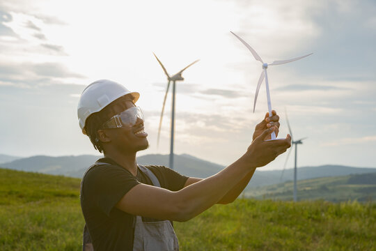 A smiling African American man in protective gear holds a miniature windmill with a wind turbine farm in the background