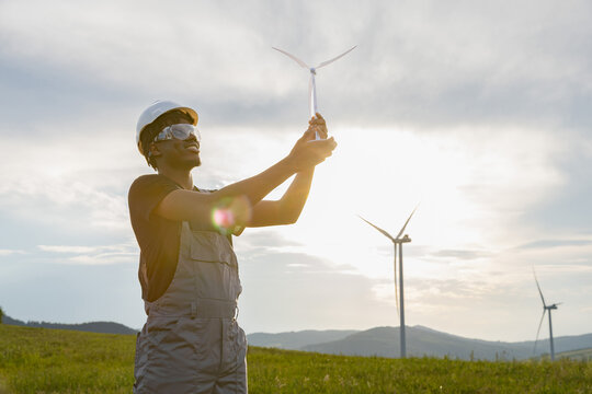 A smiling African American man in work clothes and safety gear holds a small wind turbine model against a backdrop of large wind turbines