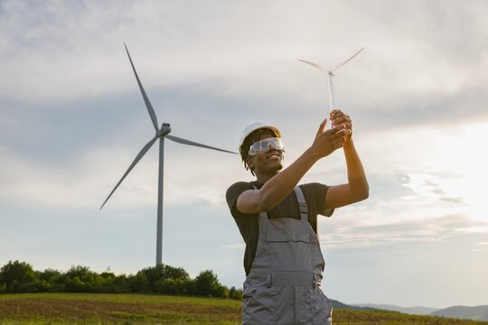 A smiling African American man in safety gear inspects a miniature wind turbine model with a large turbine in the background