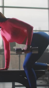 Woman performing bent-over dumbbell row on padded bench in sunlit gym, lifting metal weight toward hip while maintaining stable posture for strength