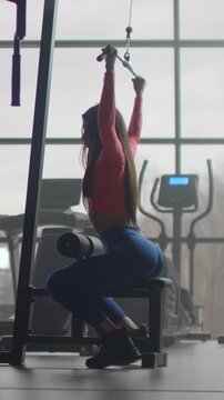 Woman seated on lat pulldown bench holding overhead cable bar behind neck in backlit gym interior showing strong controlled pull position