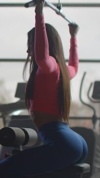 Fit woman seated at cable machine in gym, preparing lat pulldown with both hands gripping overhead bar and raised arms, strong upright posture