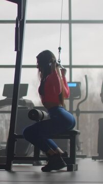 Woman seated at cable machine doing lat pulldown with arms extended overhead in backlit gym near window