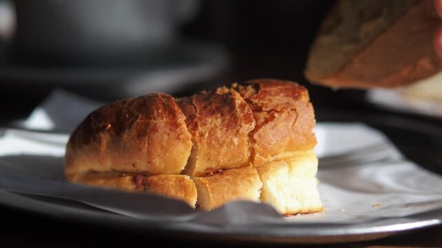 Freshly baked garlic bread roll on plate while hand picks up piece in warm morning sunlight.