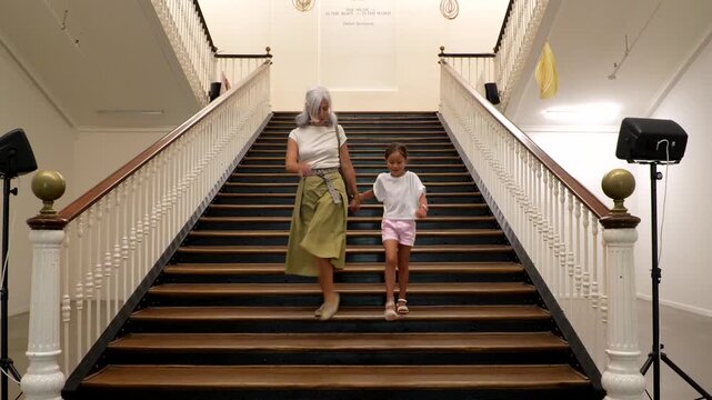 Joyful grandmother and her young granddaughter holding hands while running down a beautiful wooden staircase, smiling and having fun during a visit to a gallery or museum