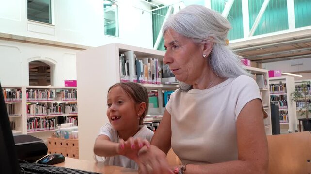 Loving grandmother with gray hair teaching her curious granddaughter how to use a computer in a public library, sharing a joyful moment of intergenerational learning and bonding